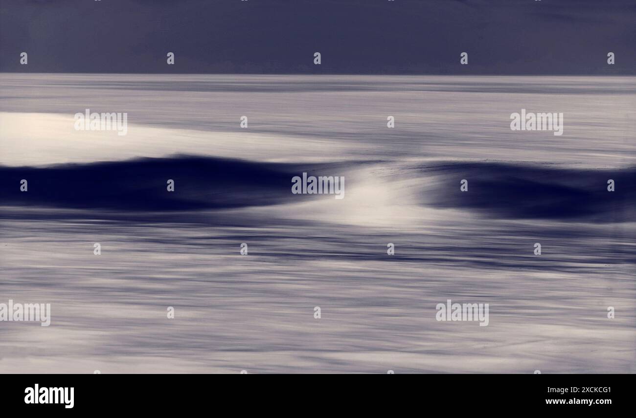 Una tranquilla scena di un'onda dolce che si infrangono su una spiaggia sabbiosa mentre il sole tramonta, che proietta un caldo bagliore sull'acqua e sulla sabbia. L'acqua è un turchese vibrante Foto Stock