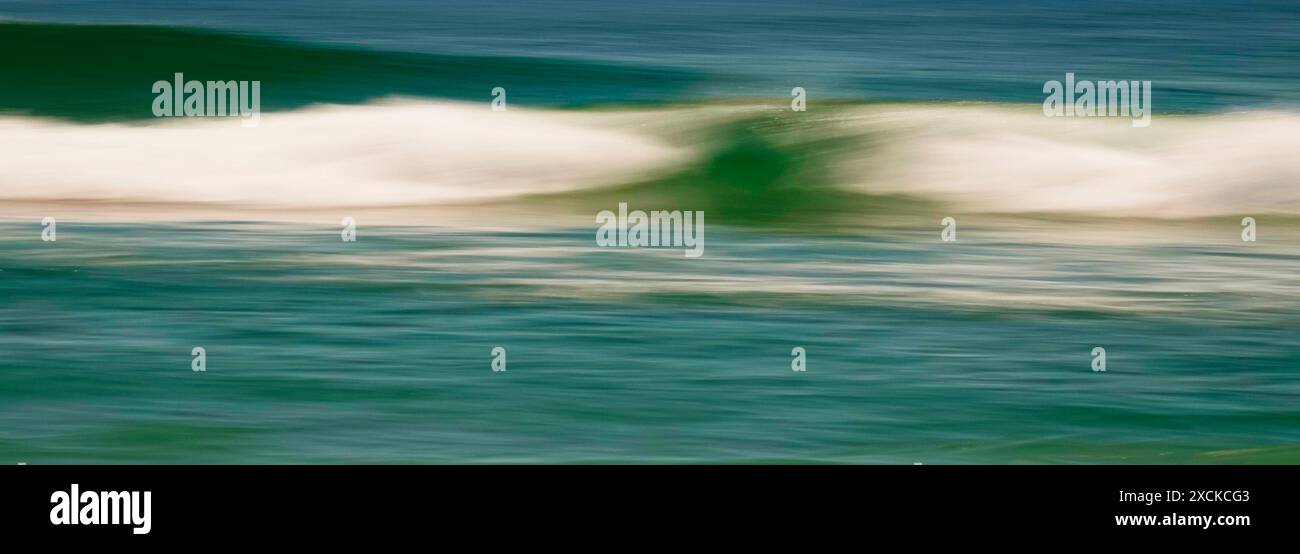 Una tranquilla scena di un'onda dolce che si infrangono su una spiaggia sabbiosa mentre il sole tramonta, che proietta un caldo bagliore sull'acqua e sulla sabbia. L'acqua è un turchese vibrante Foto Stock