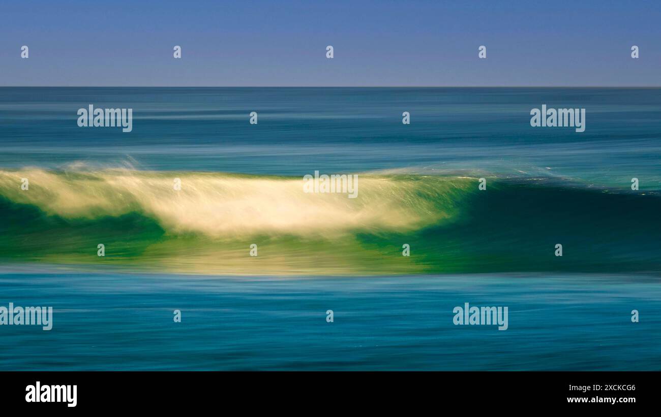 Una tranquilla scena di un'onda dolce che si infrangono su una spiaggia sabbiosa mentre il sole tramonta, che proietta un caldo bagliore sull'acqua e sulla sabbia. L'acqua è un turchese vibrante Foto Stock