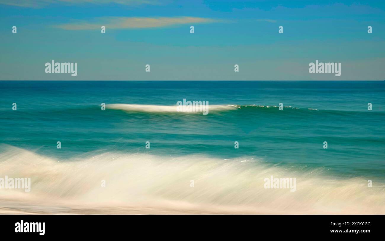 Una tranquilla scena di un'onda dolce che si infrangono su una spiaggia sabbiosa mentre il sole tramonta, che proietta un caldo bagliore sull'acqua e sulla sabbia. L'acqua è un turchese vibrante Foto Stock