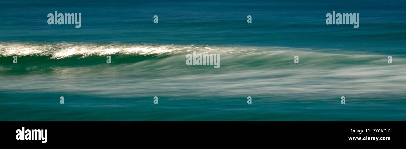 Una tranquilla scena di un'onda dolce che si infrangono su una spiaggia sabbiosa mentre il sole tramonta, che proietta un caldo bagliore sull'acqua e sulla sabbia. L'acqua è un turchese vibrante Foto Stock