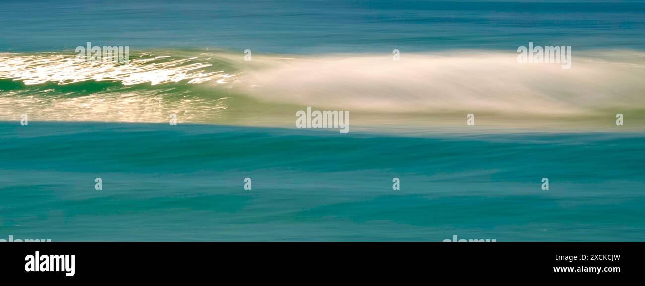 Una tranquilla scena di un'onda dolce che si infrangono su una spiaggia sabbiosa mentre il sole tramonta, che proietta un caldo bagliore sull'acqua e sulla sabbia. L'acqua è un turchese vibrante Foto Stock