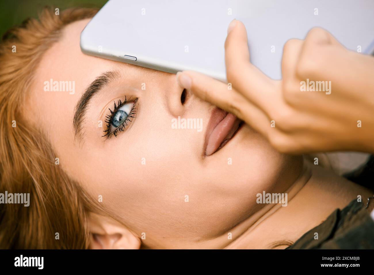 Una giovane donna con capelli biondi e occhi blu nasconde giocosamente il viso usando un tablet, creando un senso di mistero e privacy. Incarna cifre moderne Foto Stock