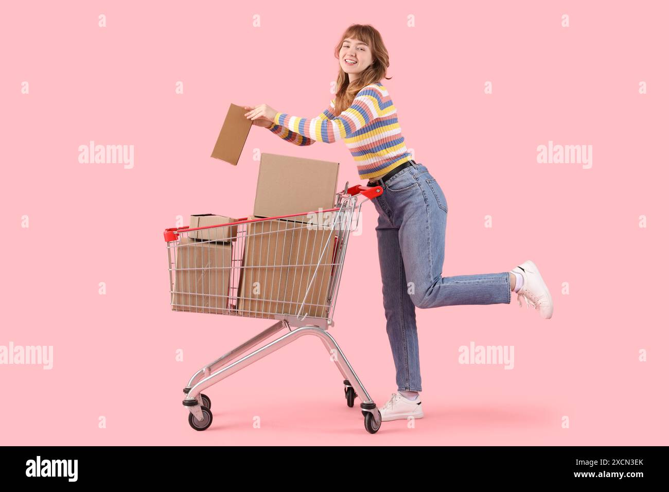 Giovane donna con carrello pieno di scatole di cartone su sfondo rosa Foto Stock