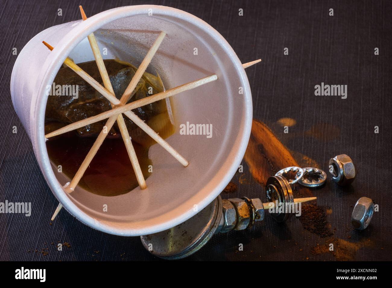 Tazza monouso in polistirene bianco con sacchetto per il tè e tè disposti su sfondo scuro con dadi e bulloni arrugginiti. Foto Stock