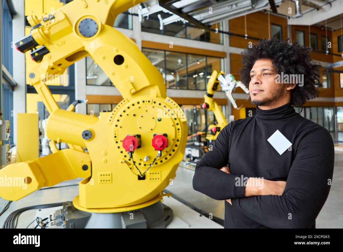 Un ingegnere in piedi con sicurezza accanto a un grande braccio robotico giallo in un ambiente industriale, che rappresenta lo sviluppo dell'automazione, la programmazione, un Foto Stock