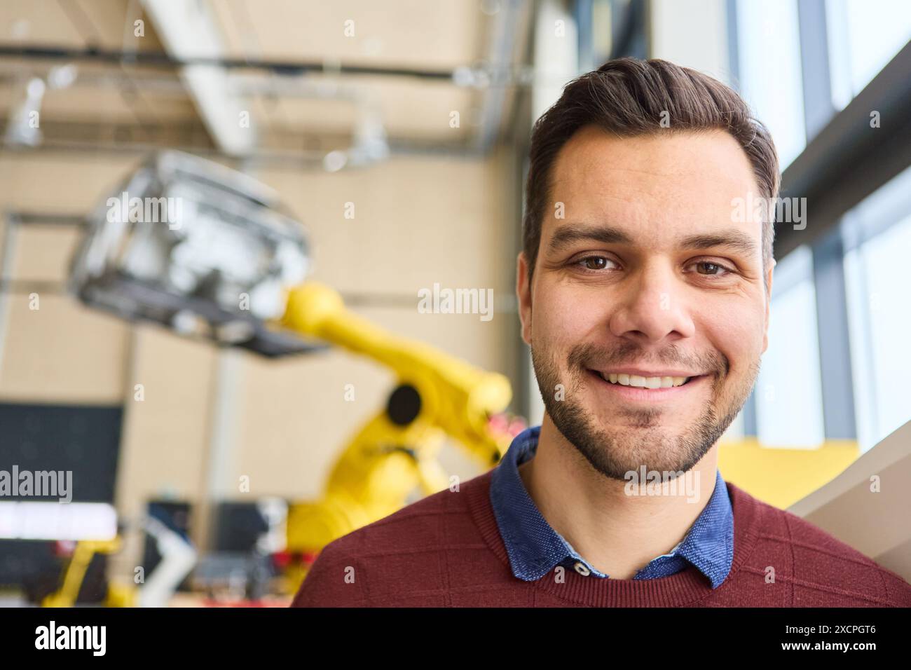 Un ingegnere felice sorridente in un moderno laboratorio di robotica con un braccio robotico industriale sullo sfondo, che rappresenta innovazione e tecnologia. Foto Stock