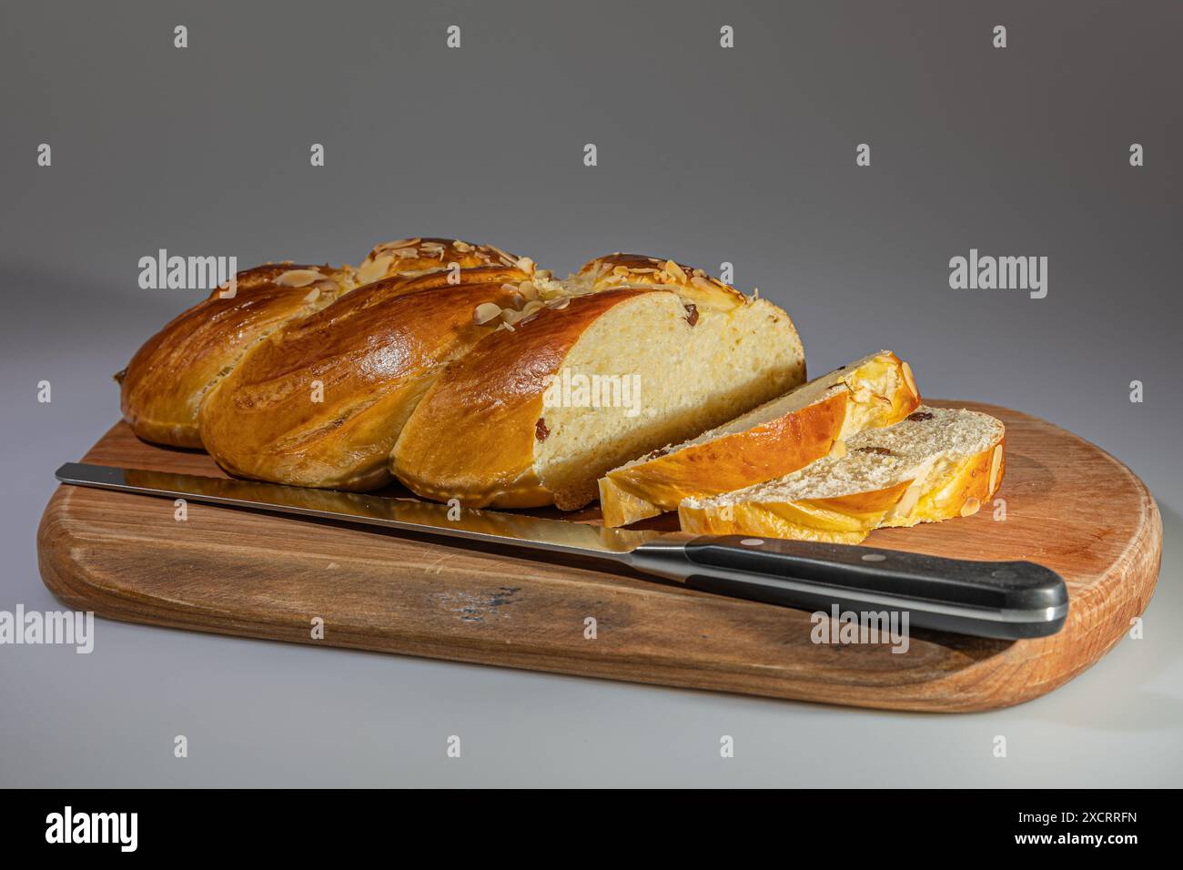 Un pane intrecciato appena sfornato, ricoperto di mandorle e ripieno di uvetta, è parzialmente tagliato a fette e esposto su un tagliere di legno. Il brea Foto Stock