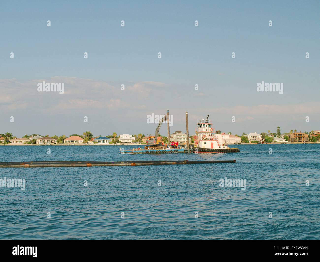 Ampia vista sul canale della griglia Pass-A. Draga chiatta, gru e tubi in acqua blu. Nuvole ed edifici bianchi del cielo blu Foto Stock