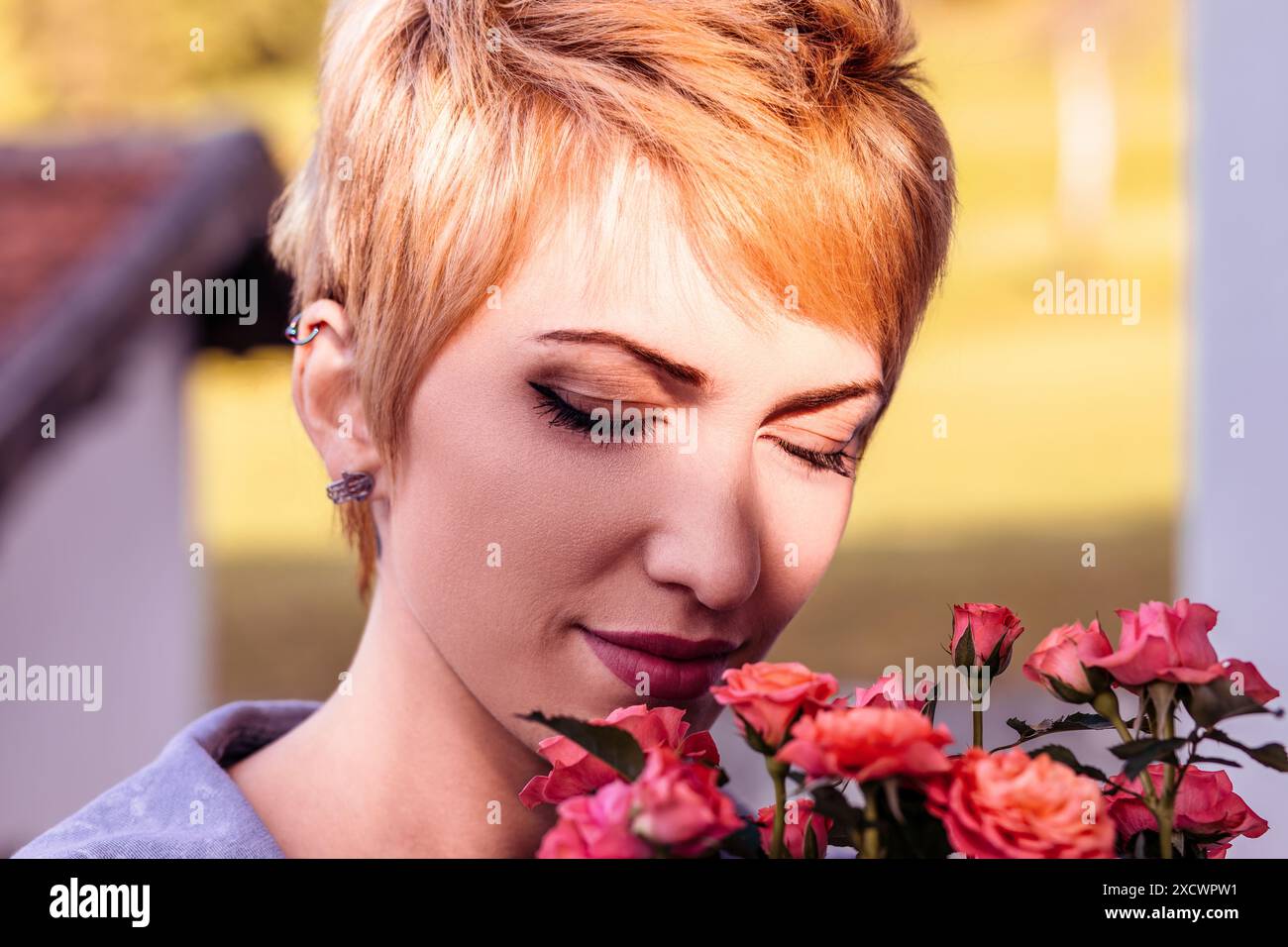 Una giovane donna con i capelli biondi corti e un naso che piercing chiude gli occhi e profuma di rose rosa all'aperto Foto Stock