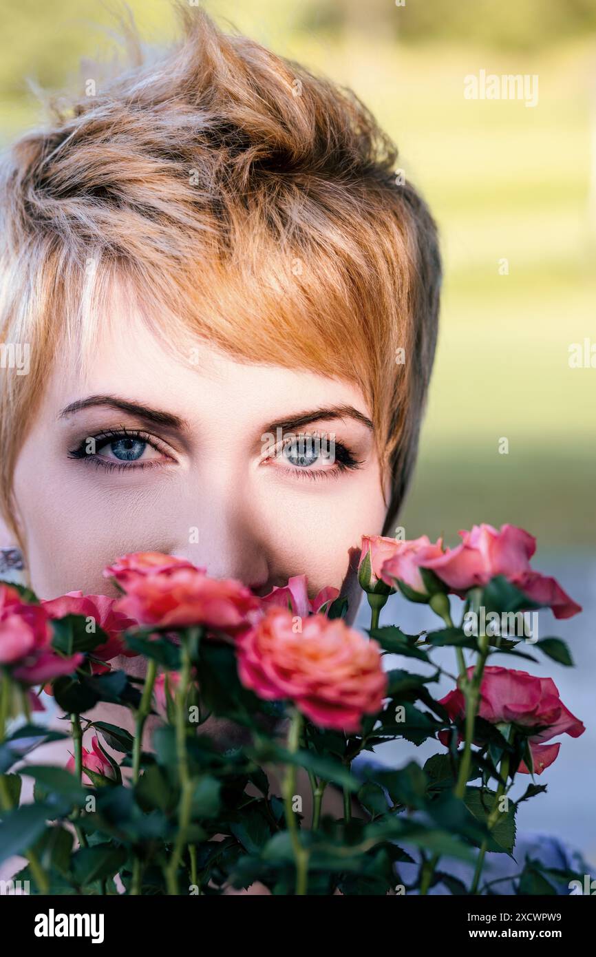 La giovane donna dai capelli biondi corti copre parte del viso con un bouquet di rose rosa, godendosi la loro fragranza e bellezza in una giornata di sole Foto Stock