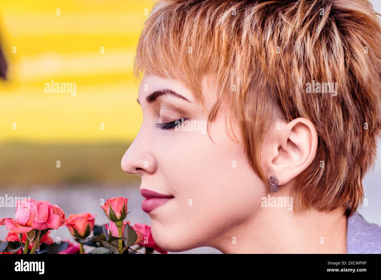 Giovane donna dai capelli biondi corti che si gode il profumo delle rose rosa e gli occhi chiusi in tranquillità. All'aperto in un giardino, trasuda pace e felicità Foto Stock