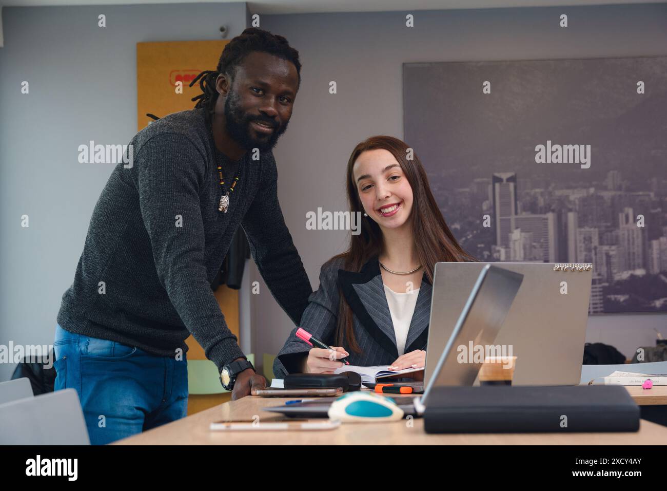 Uomo adulto con dreadlock in piedi accanto alla sua collega donna seduta con un laptop e che guarda la fotocamera all'interno di coworking. L'uomo indossa una swe grigia Foto Stock