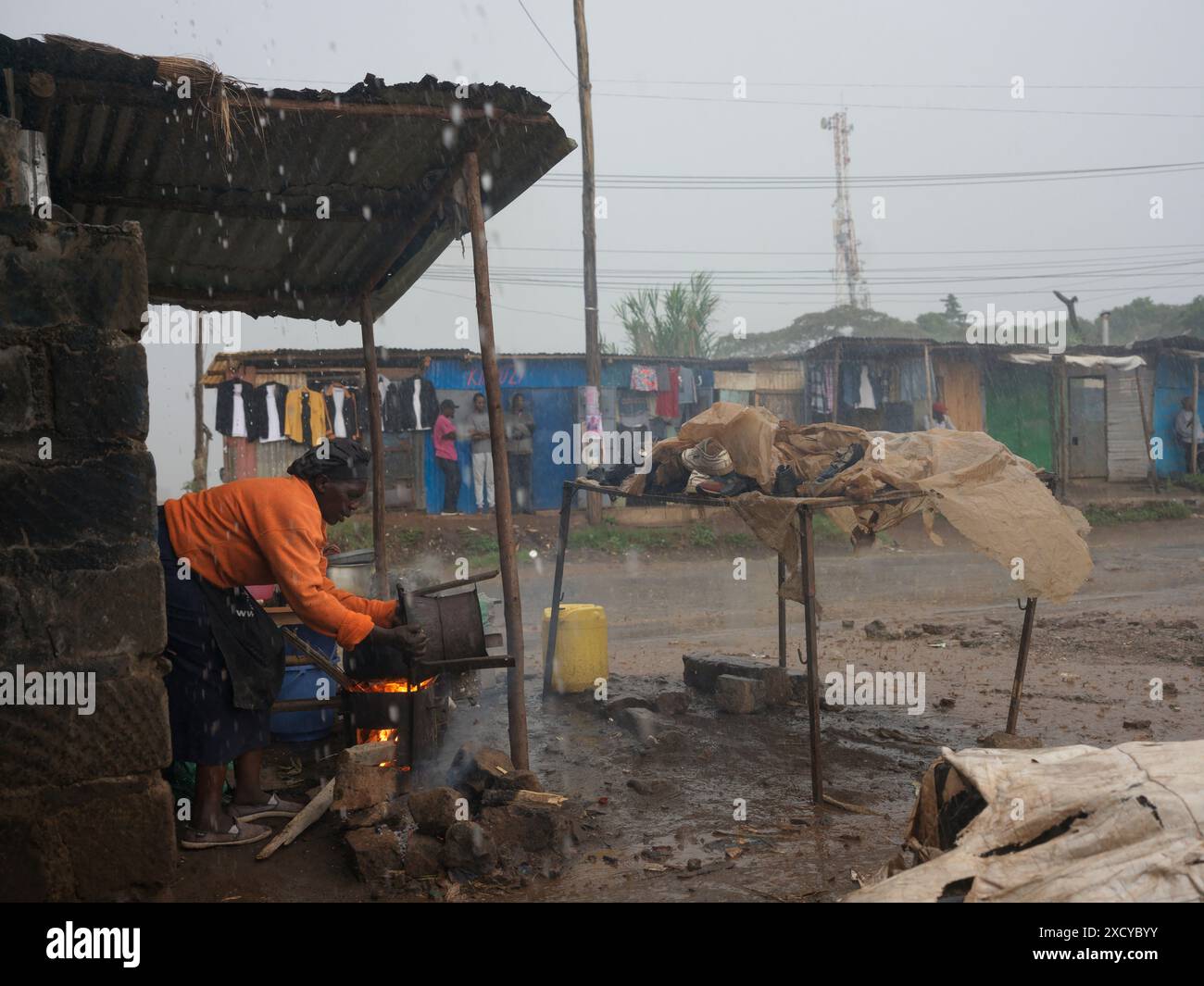 Donna al riparo dalla pioggia usando una stufa a legna o a carbone per cucinare un pasto nella baraccopoli di Kibera, Nairobi, Kenya Foto Stock