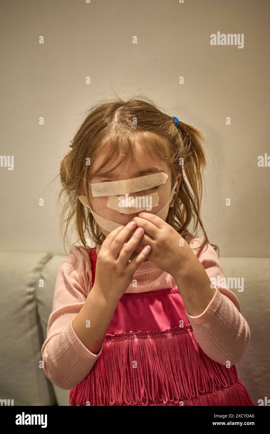La ragazza in abito fucsia ha 3 strisce di nastro di carta attaccate sul viso, coprendo gli occhi, il naso e la bocca mentre tocca la bocca con le mani. Lei è forc Foto Stock