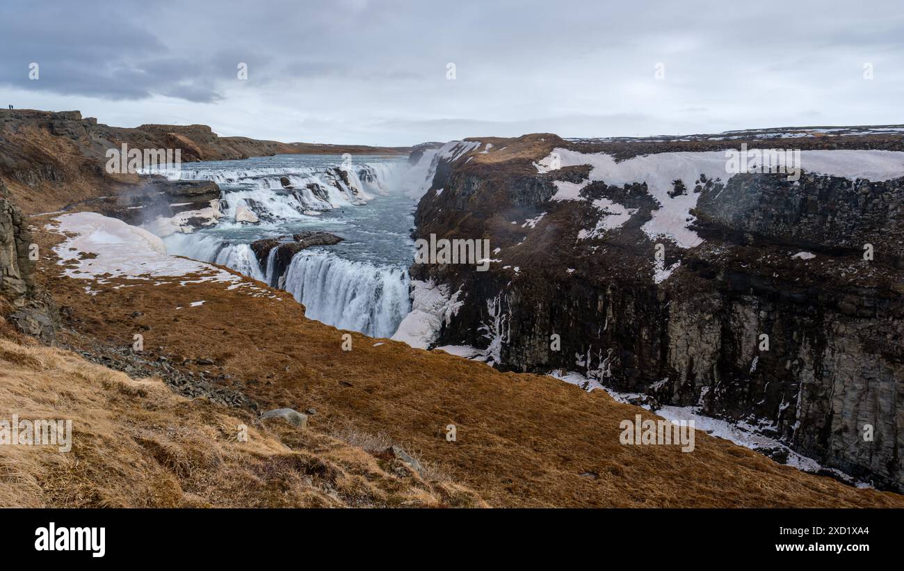 Una vista mozzafiato della cascata Gullfoss in Islanda, circondata da scogliere innevate e terreno accidentato sotto un cielo nuvoloso. Foto Stock