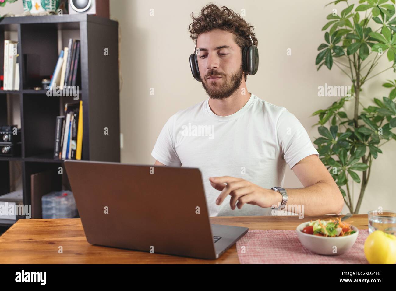 Un uomo che lavora da casa con un computer portatile, indossa le cuffie e prepara un'insalata salutare. Ideale per contenuti su lavoro remoto, tecnologia e sane lif Foto Stock