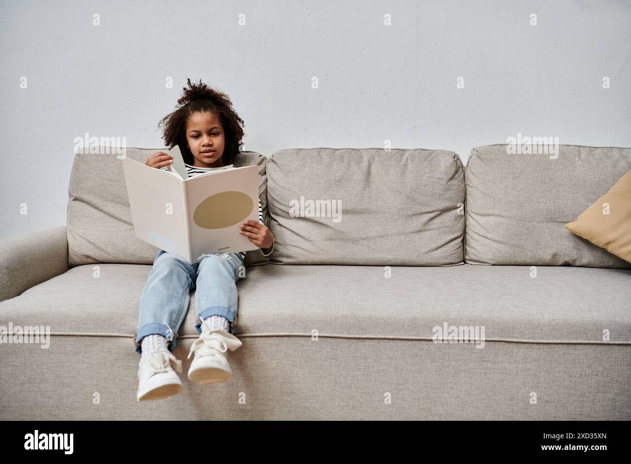 Una bambina di origini afroamericane siede su un comodo divano, infilata in un libro Foto Stock