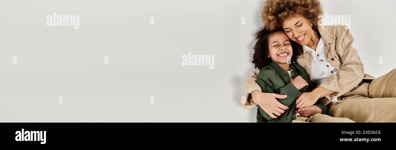 Una madre riccia afro-americana abbraccia amorevolmente sua figlia, entrambe in abiti eleganti, su uno sfondo grigio. Foto Stock