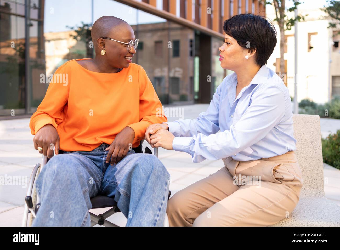 Una donna nera sorridente su una sedia a rotelle impegnata in una discussione amichevole e di sostegno con una donna latina seduta accanto a lei, entrambi all'aperto in una città Foto Stock