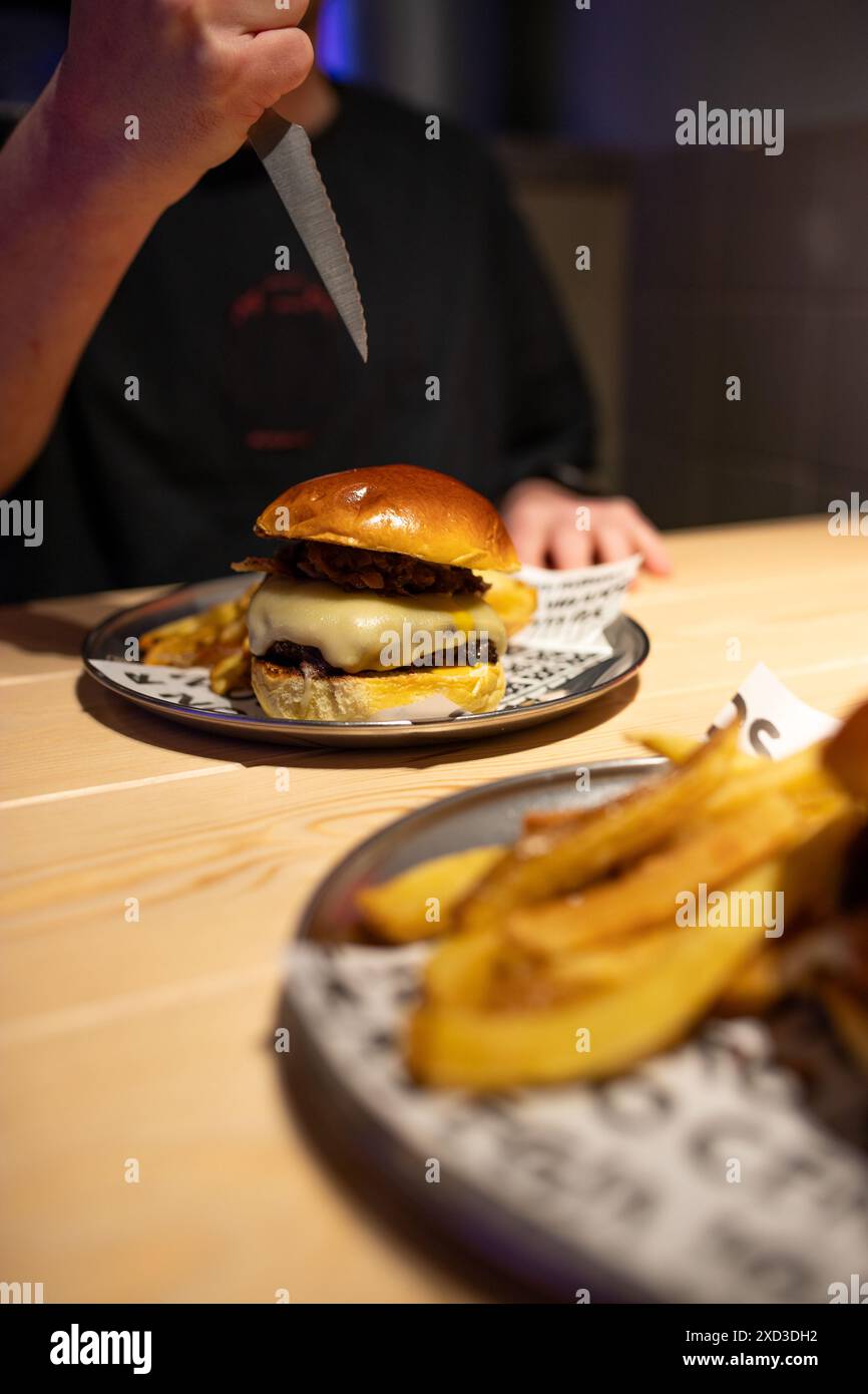 Una persona con una camicia scura sta per affettare un succoso hamburger gourmet impilato servito su un piatto con un lato di patatine fritte dorate in un ristorante moderno Foto Stock