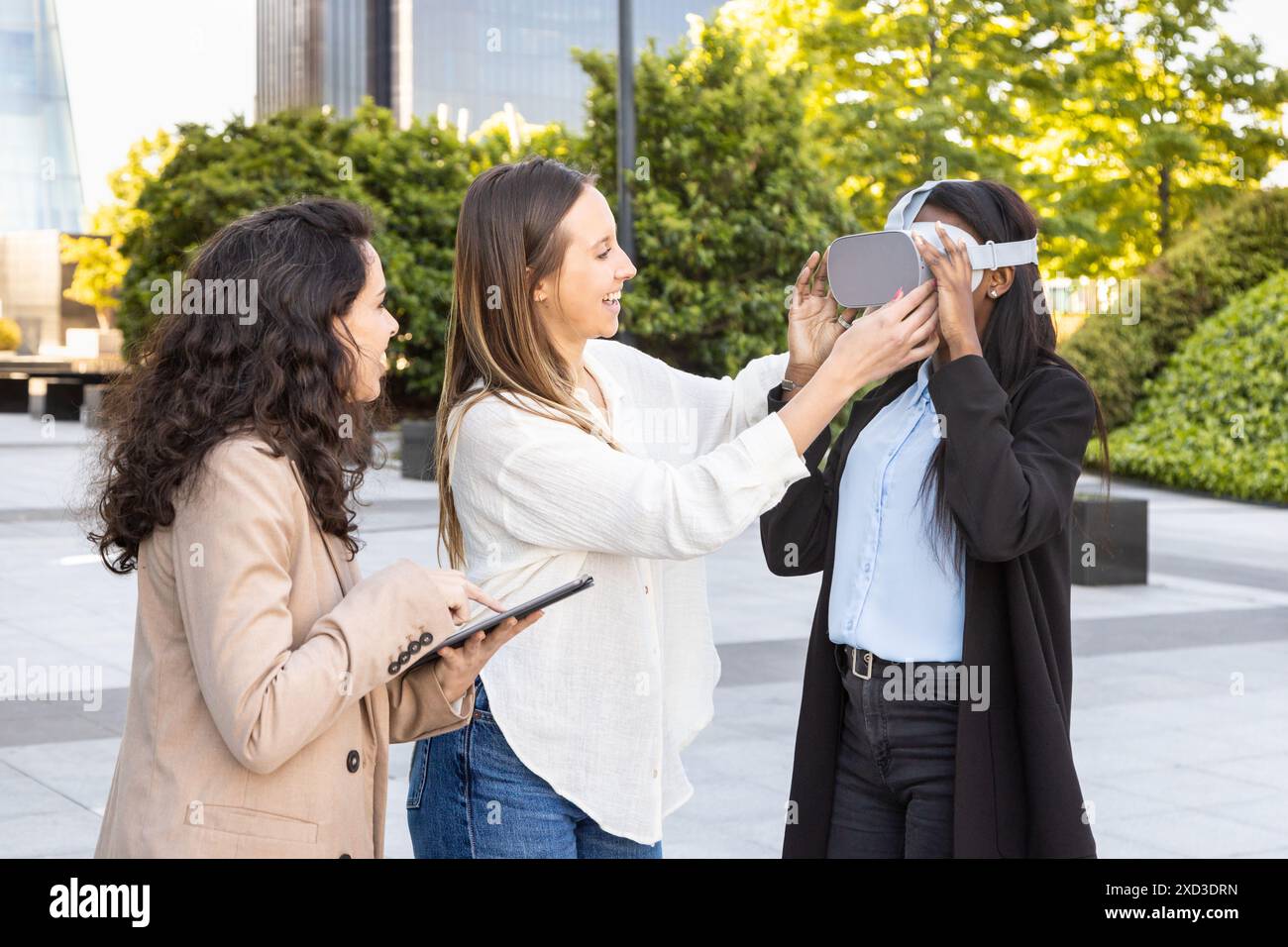Tre donne professionali con background diversi collaborano all'aperto, utilizzando un visore per la realtà virtuale in un parco urbano con edifici moderni aro Foto Stock