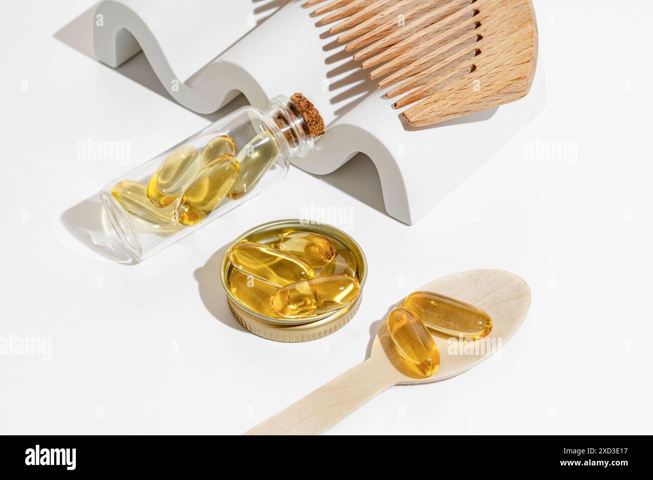 I contenitori in vetro trasparente e un cucchiaio di legno contengono capsule Omega 3 dorate, posizionate su uno sfondo bianco per un look pulito e moderno Foto Stock