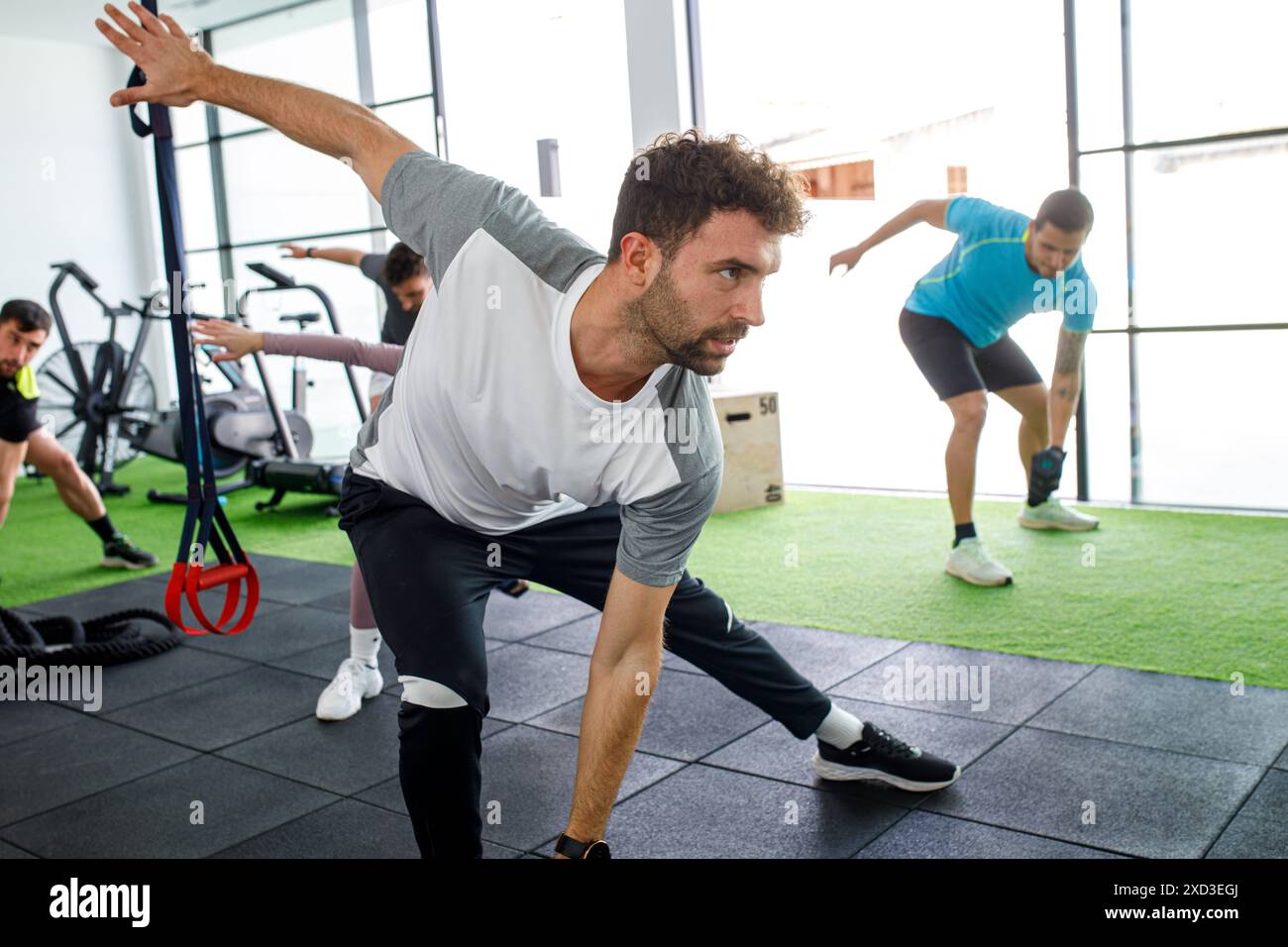 Immagine che raffigura giovani adulti che partecipano ad esercizi di stretching di gruppo in una palestra contemporanea, circondata da attrezzature fitness e grandi finestre Foto Stock