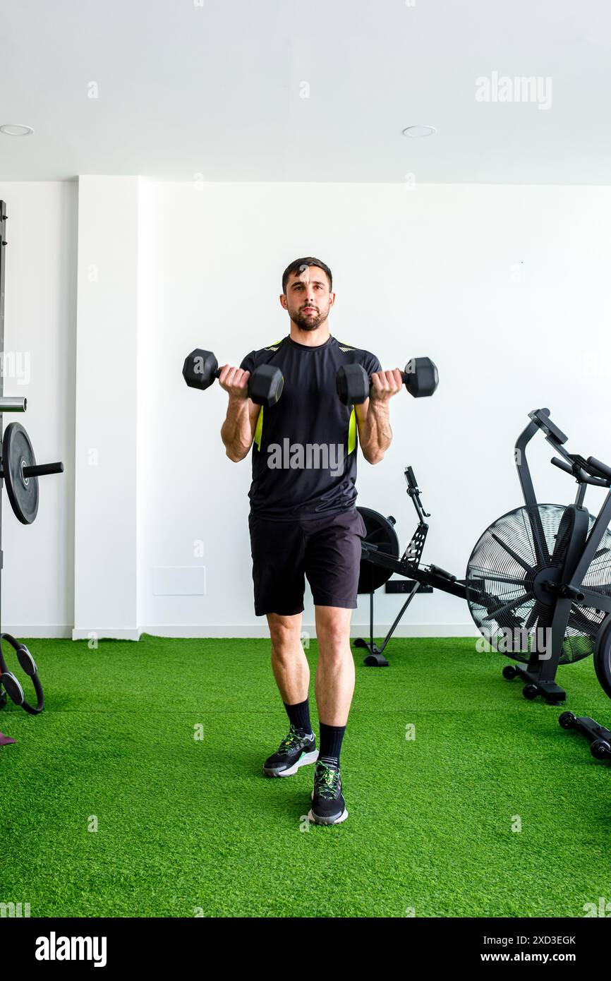 Un giovane attento solleva pesanti manubri all'interno di una palestra spaziosa e ben illuminata con pareti bianche, pavimento in erba verde e varie attrezzature per esercizi Foto Stock Un giovane attento solleva pesanti manubri all'interno di una palestra spaziosa e ben illuminata con pareti bianche, pavimento in erba verde e varie attrezzature per esercizi Foto Stock