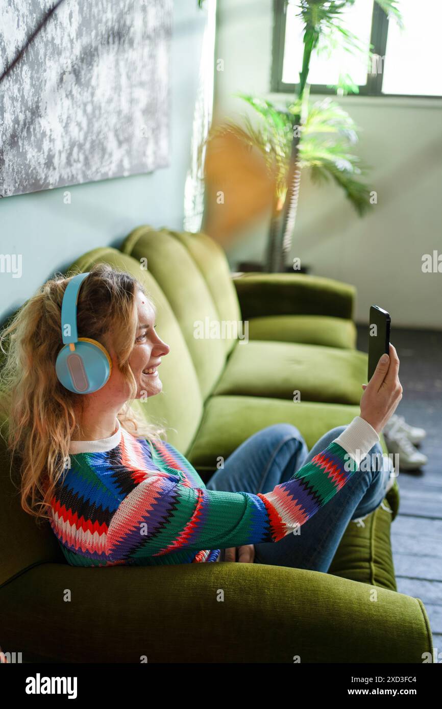 Una giovane donna allegra si gode una pausa su un divano verde, ascoltando musica con le cuffie e utilizzando lo smartphone in una stanza piena di luce Foto Stock