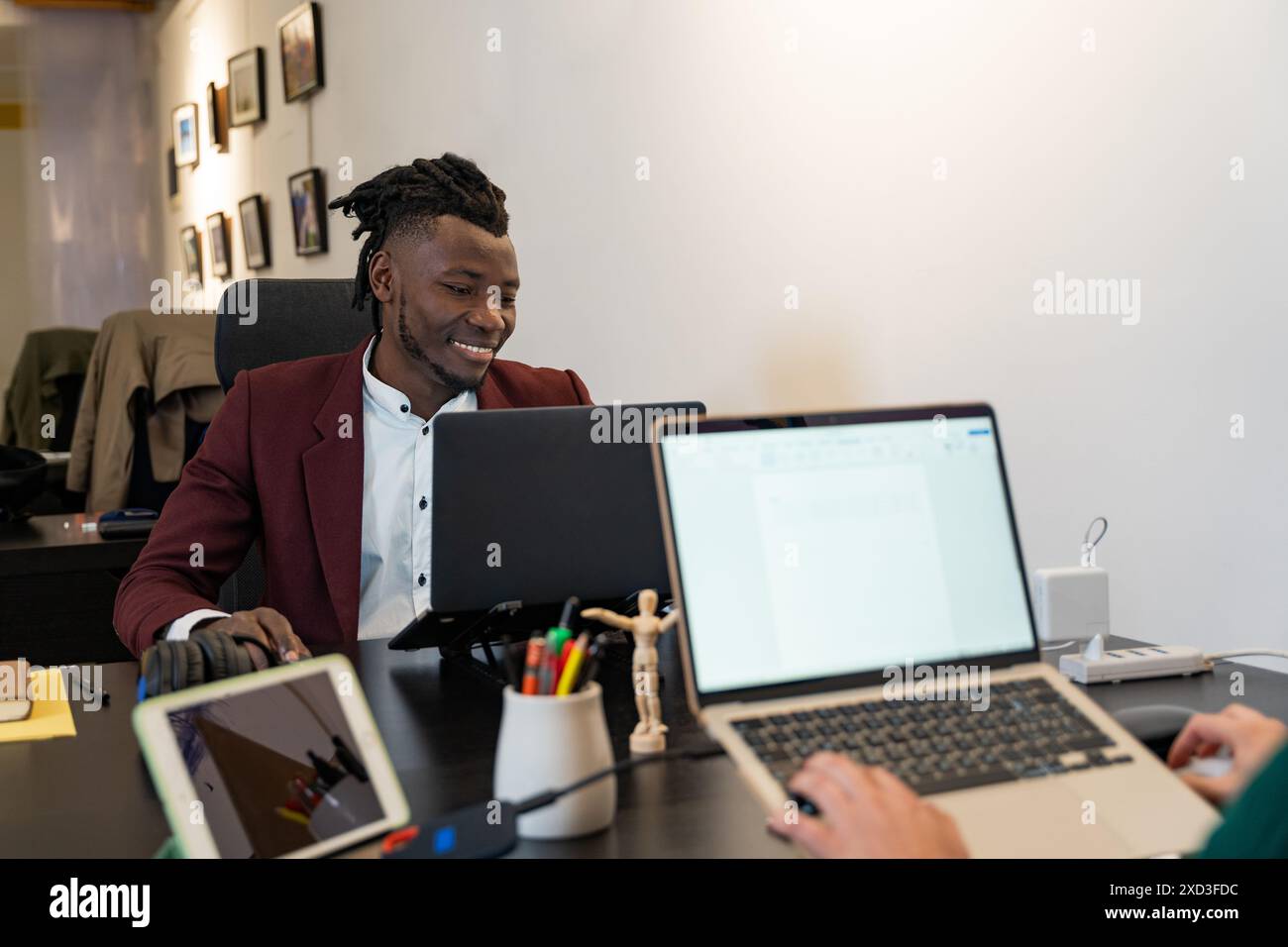 Un giovane uomo d'affari allegro in un elegante blazer color marrone lavora con attenzione sul suo notebook, interagendo con un collega di un ufficio moderno e ben attrezzato Foto Stock