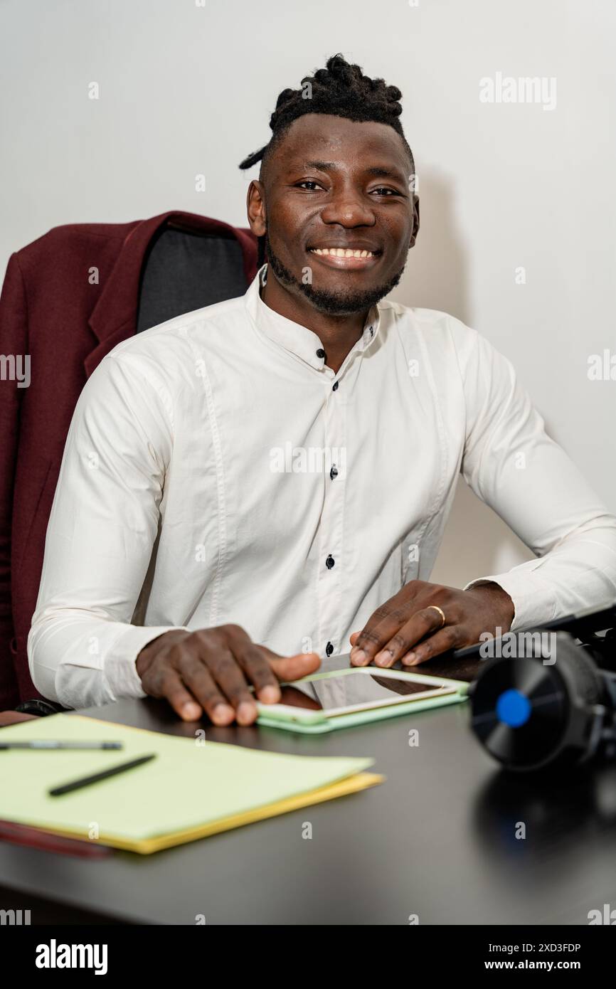 Un giovane allegro con una camicia bianca si siede alla scrivania dell'ufficio, sorridendo calorosamente mentre si occupa di attività legate al lavoro è circondato da ess dell'ufficio Foto Stock