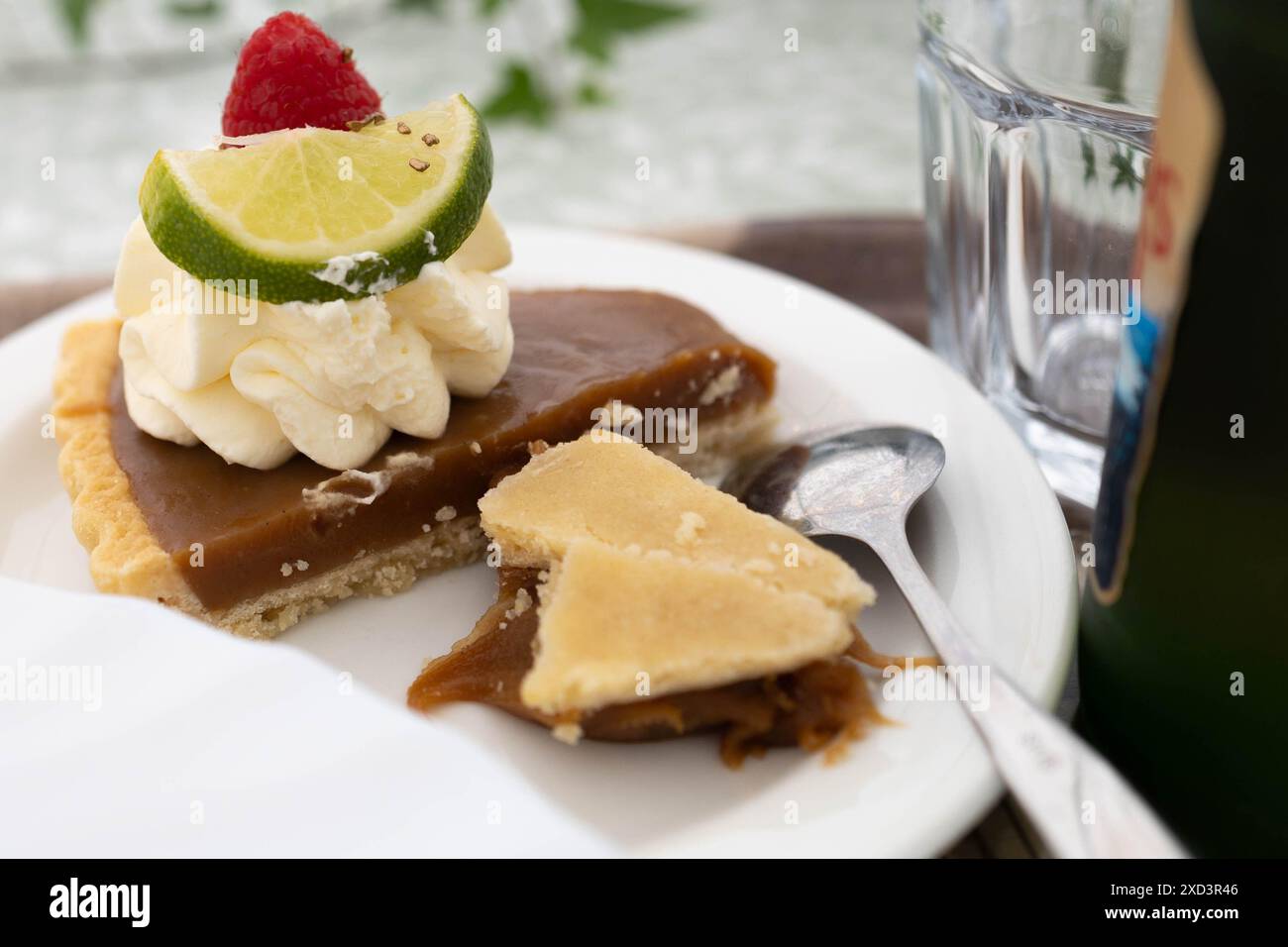 Primo piano di una fetta di crostata al caramello condita con panna montata, lime e lampone, servita su un piatto bianco con un cucchiaio. Foto Stock