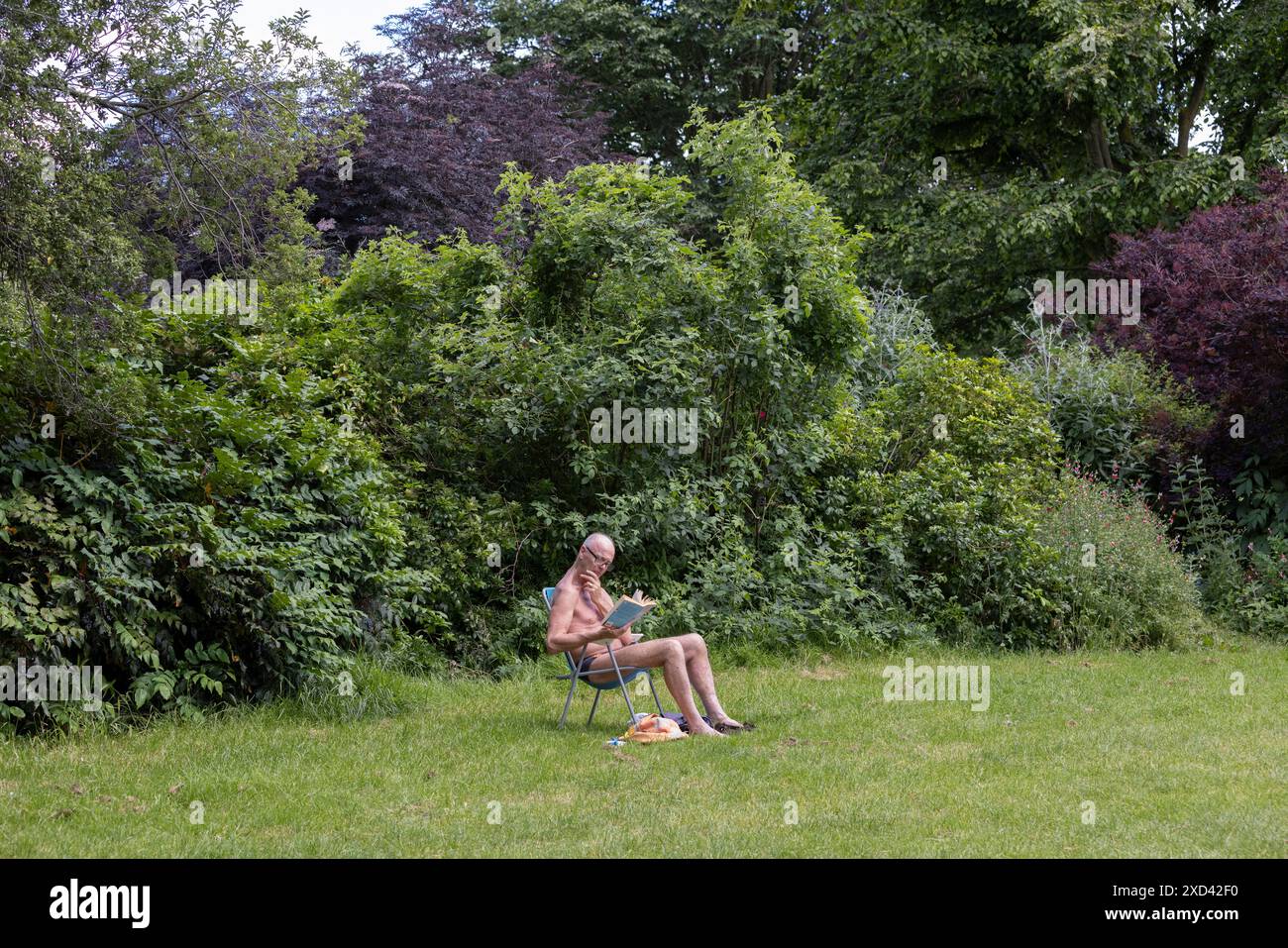 Un uomo siede a leggere su una sedia a sdraio a prendere il sole nei Kensington Palace Gardens durante una mini ondata di calore a Londra, Inghilterra, Regno Unito Foto Stock