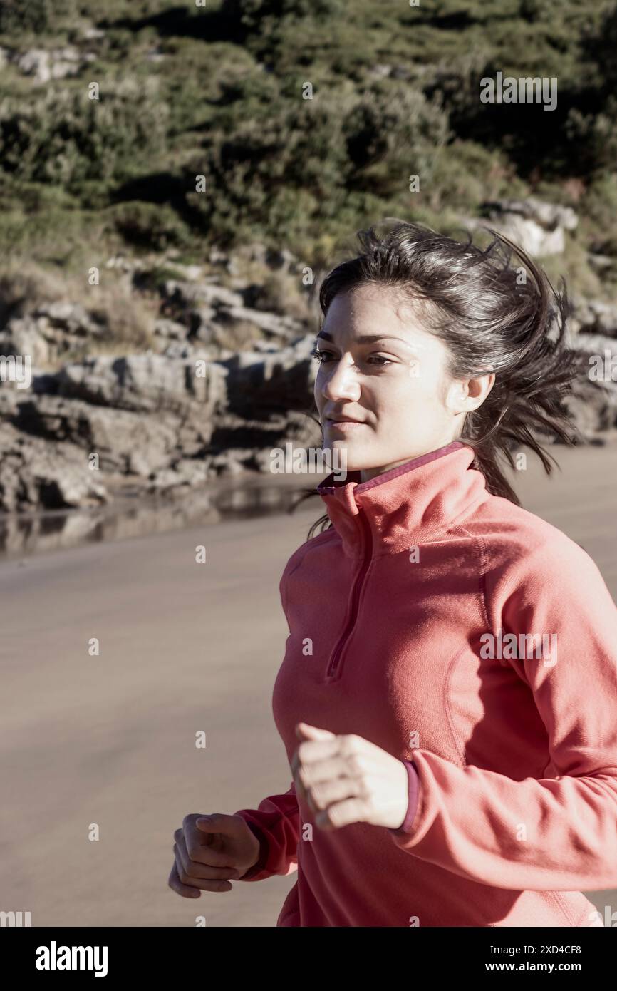 Giovane donna che fa jogging sulla spiaggia con giacca rosa. Fitness e fotografia di stile di vita sano. Concetto di esercizio e benessere Foto Stock