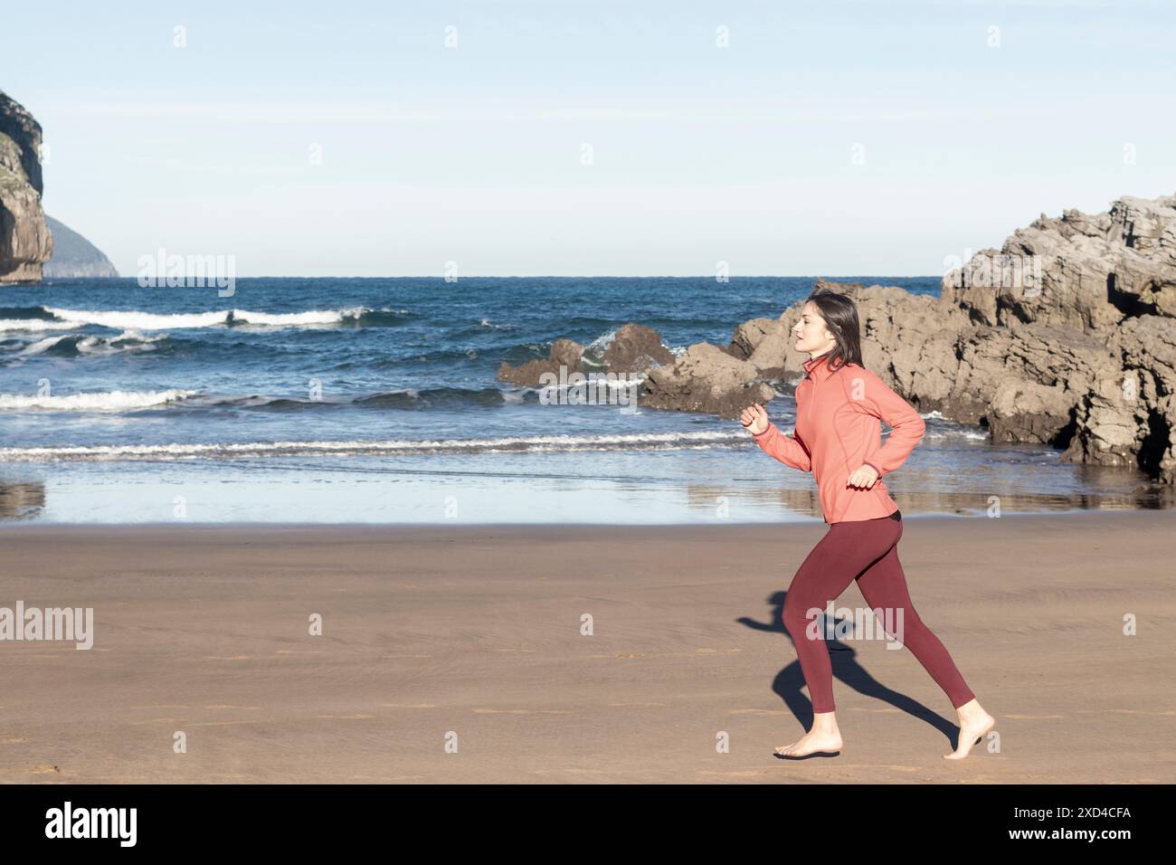 Giovane donna che fa jogging a piedi nudi sulla spiaggia con giacca rosa e leggings rossi. fitness e fotografia di uno stile di vita sano. Concetto di esercizio e benessere Foto Stock