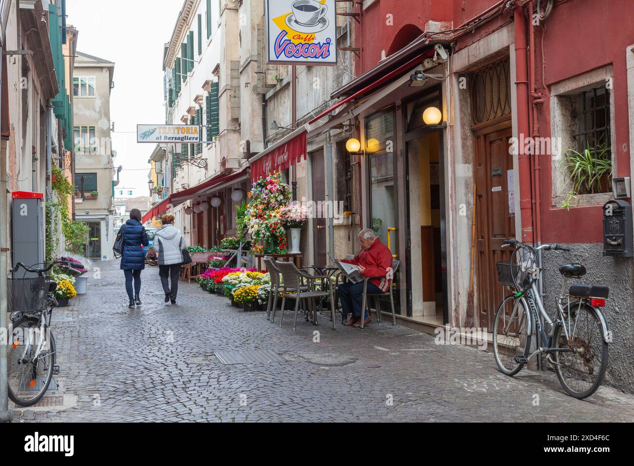 Vita quotidiana a Chioggia con veneziani locali in una stretta strada acciottolata costeggiata da trattorie e uomo seduto fuori dal caffè Ducale, Venezia, Italia Foto Stock