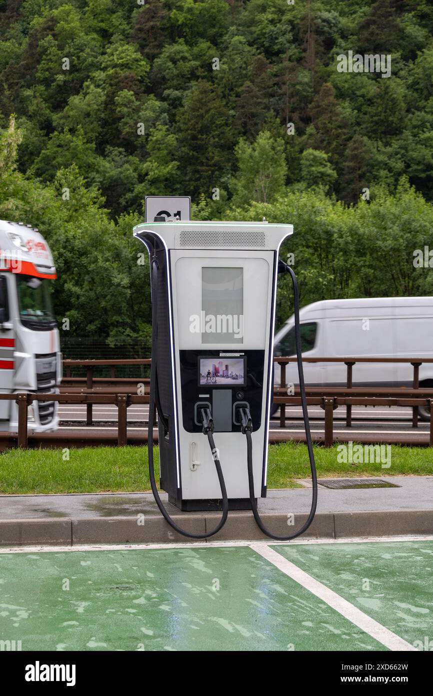 n stazione di ricarica per veicoli elettrici sull'autostrada A22 a Sudtirol, con traffico di passaggio e vegetazione lussureggiante sullo sfondo. Foto Stock