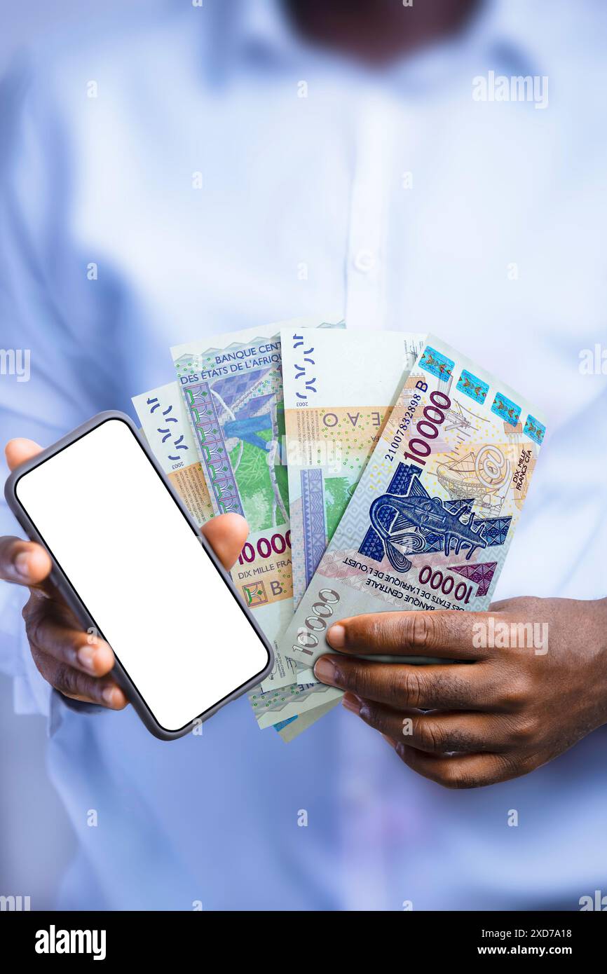 Persona nera che tiene in mano le note del franco CFA dell'Africa occidentale e mostra il telefono con schermo vuoto alla fotocamera. Mano che tiene banconote in valuta dell'Africa occidentale Foto Stock