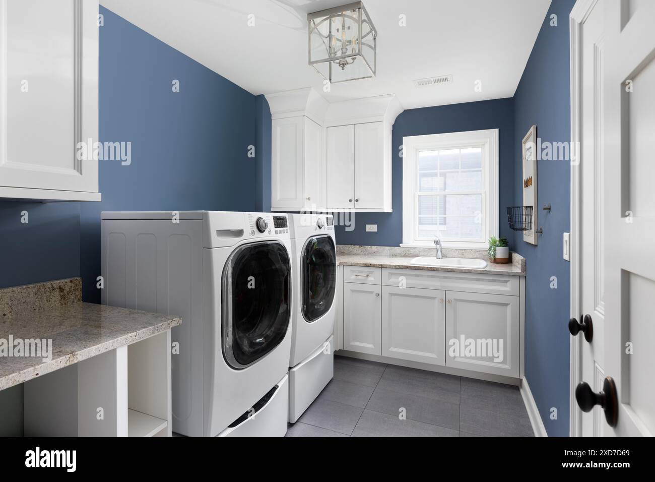 Una lavanderia con elettrodomestici e armadietti bianchi, un pavimento piastrellato grigio, un lavandino su un ripiano in granito e un apparecchio di illuminazione color argento appeso sopra. Foto Stock