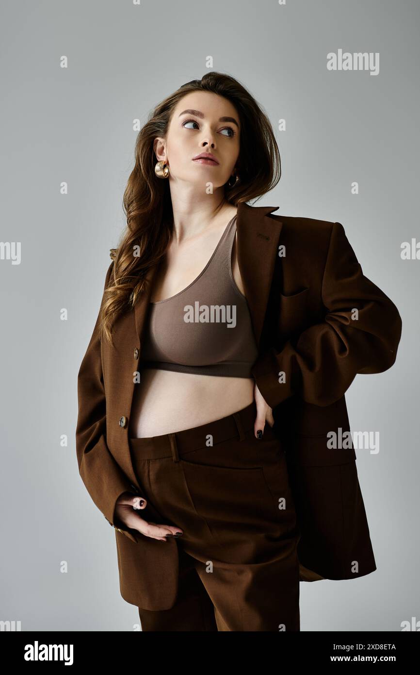 Una giovane donna incinta con un abito marrone e un blazer che si fa sentire sicura su uno sfondo grigio. Foto Stock