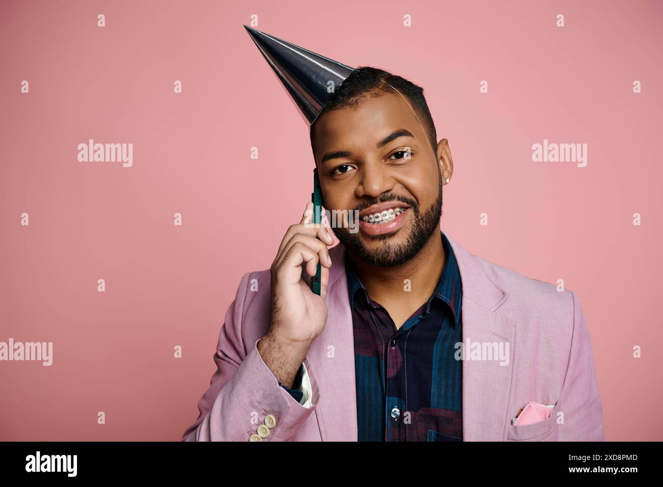 Il giovane afroamericano con l'apparecchio parla felicemente al cellulare indossando un cappello da festa sullo sfondo rosa. Foto Stock