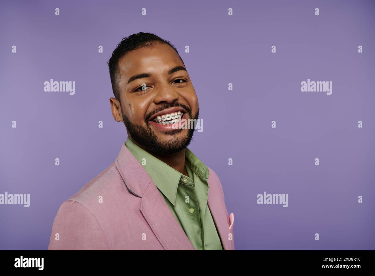 Giovane afroamericano sorridente con un elegante abito rosa e una camicia verde su un vivace sfondo viola. Foto Stock