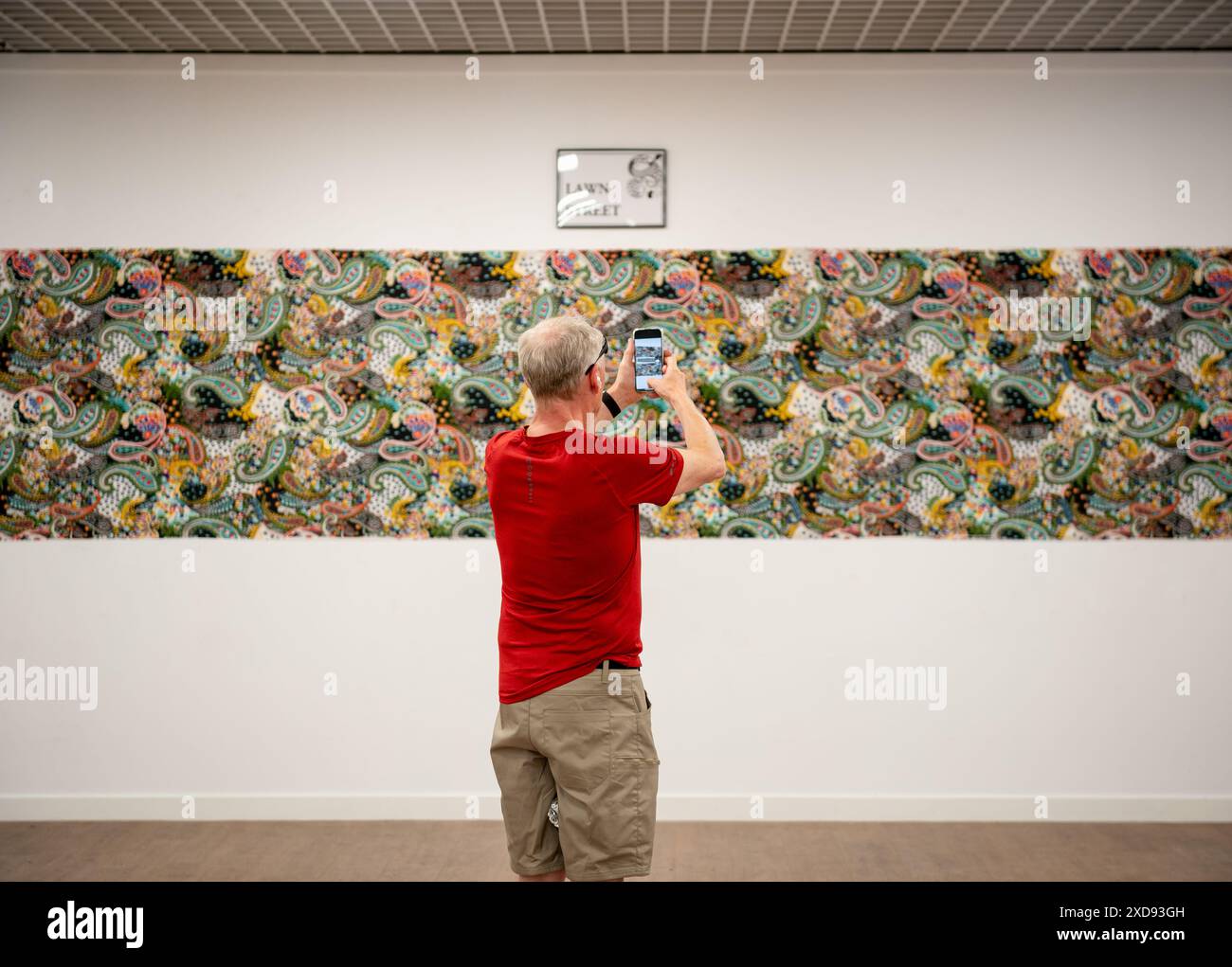 Un uomo con una maglietta rossa si trova di fronte a una lunga opera d'arte rettangolare con Paisley Pattern che scatta una foto con il suo telefono cellulare Foto Stock