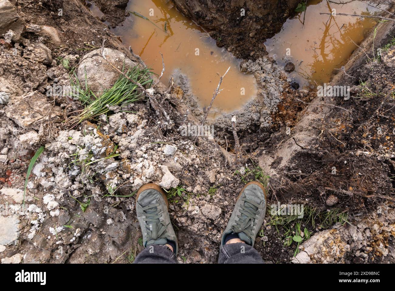 Piedi di un uomo in piedi all'angolo di una profonda fossa di fondamenta in un terreno sporco, vista in prima persona Foto Stock