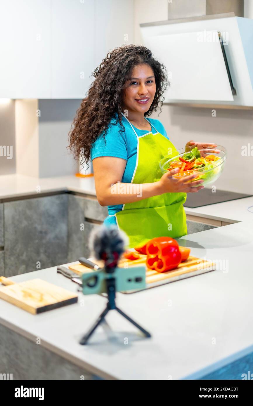 Foto verticale di una graziosa ragazza latina che mostra l'insalata alla macchina fotografica durante il tutorial online da casa Foto Stock