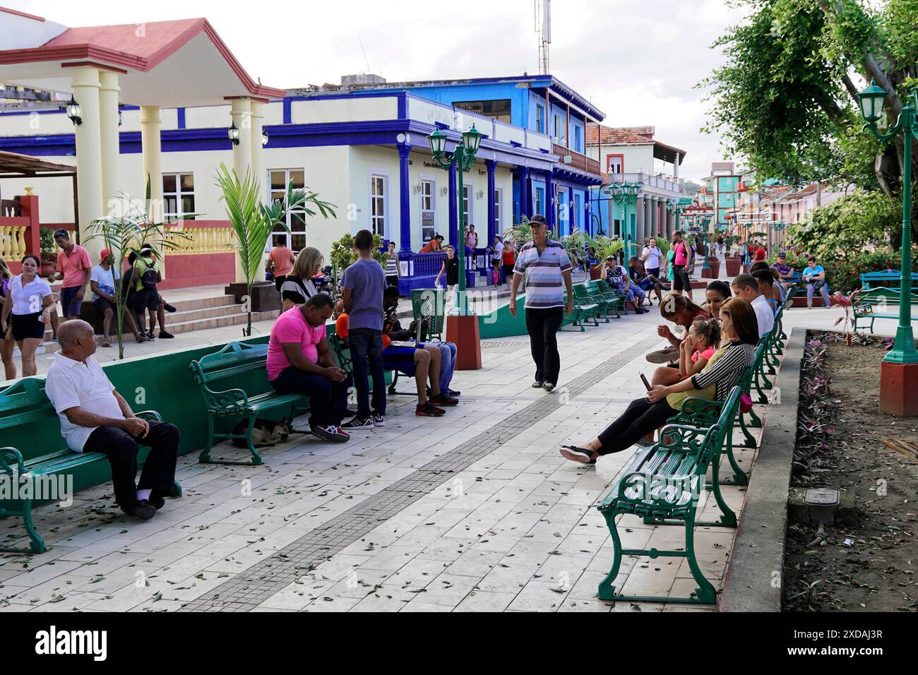 Baracoa, Cuba, America centrale, diverse persone sedute sulle panchine del parco lungo un passaggio pedonale con edifici e alberi, Cuba Foto Stock