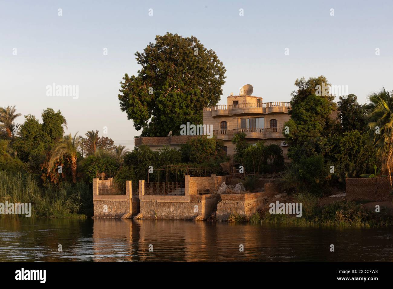 Una casa egiziana borghese in pietra, progettata da un architetto, e il suo giardino paesaggistico sulle rive del fiume Nilo vicino a Esna, risplende albicocca all'alba. Foto Stock