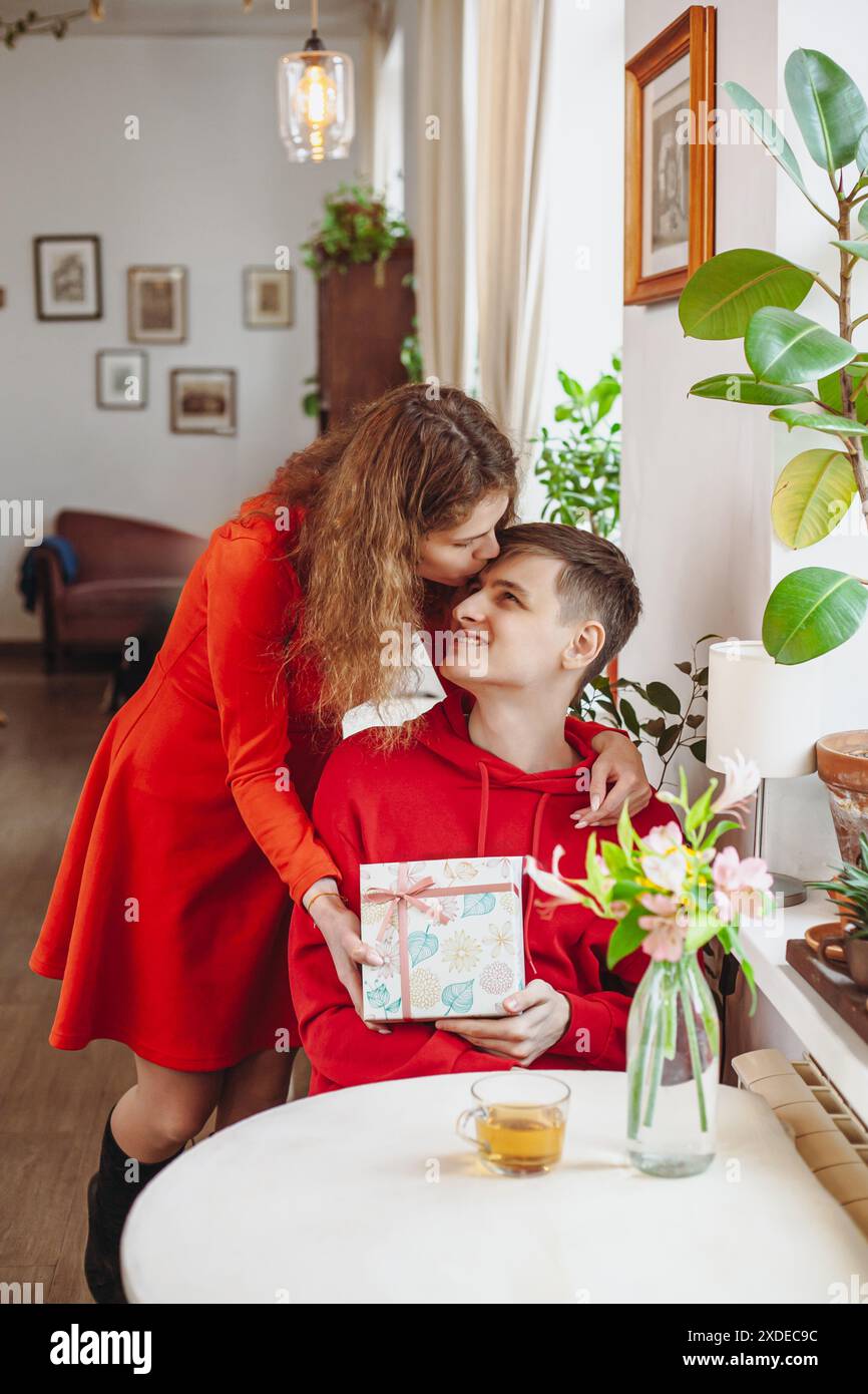 Una donna con un vestito rosso bacia un uomo sulla fronte mentre gli presenta una confezione regalo. Sono seduti ad un tavolo in un Cafe' con un bicchiere di te' e. Foto Stock