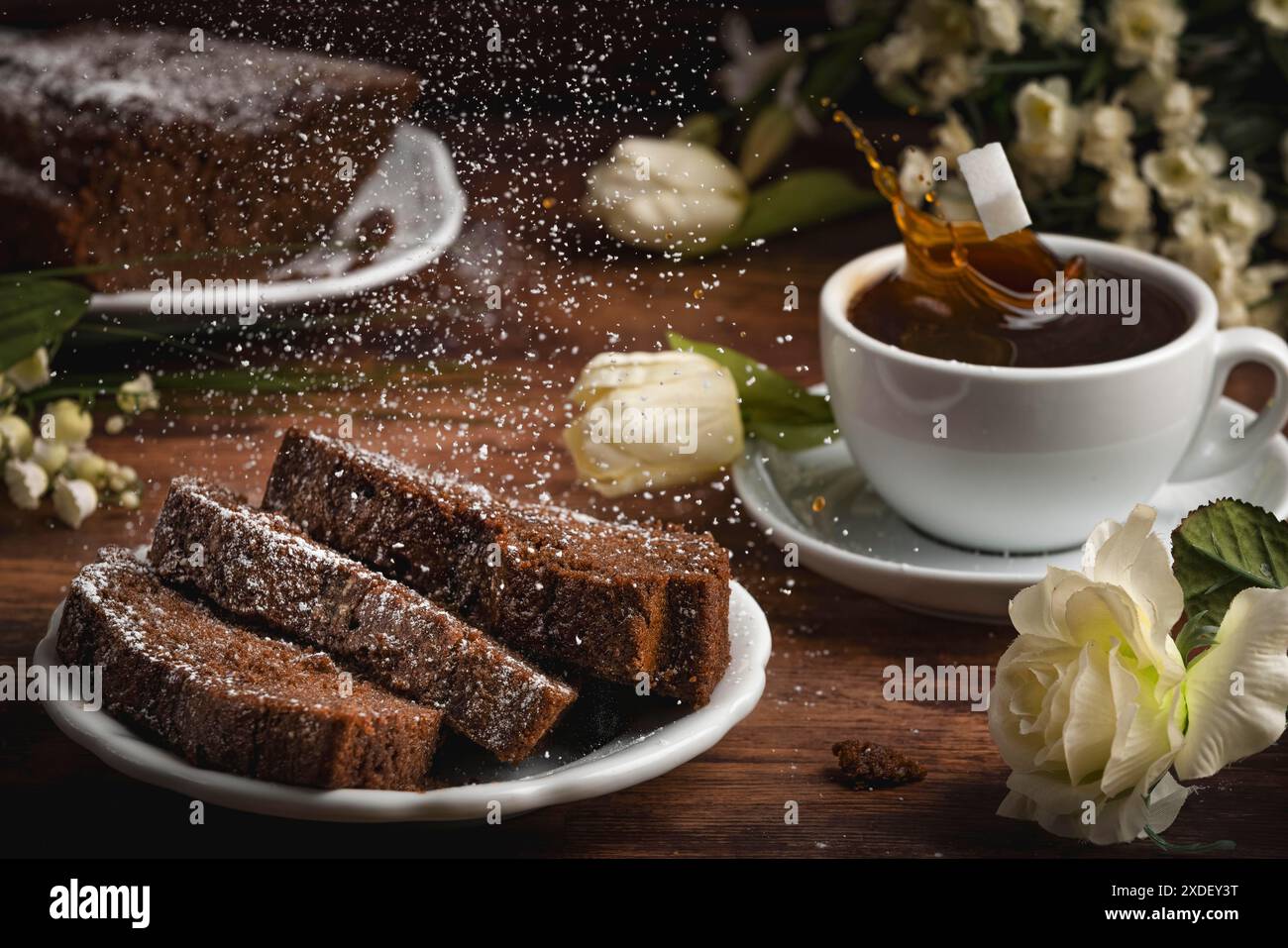 Una tazza di caffè, una torta con zucchero a velo e gocce di caffè spruzzate, una disposizione floreale su un tavolo di legno Foto Stock